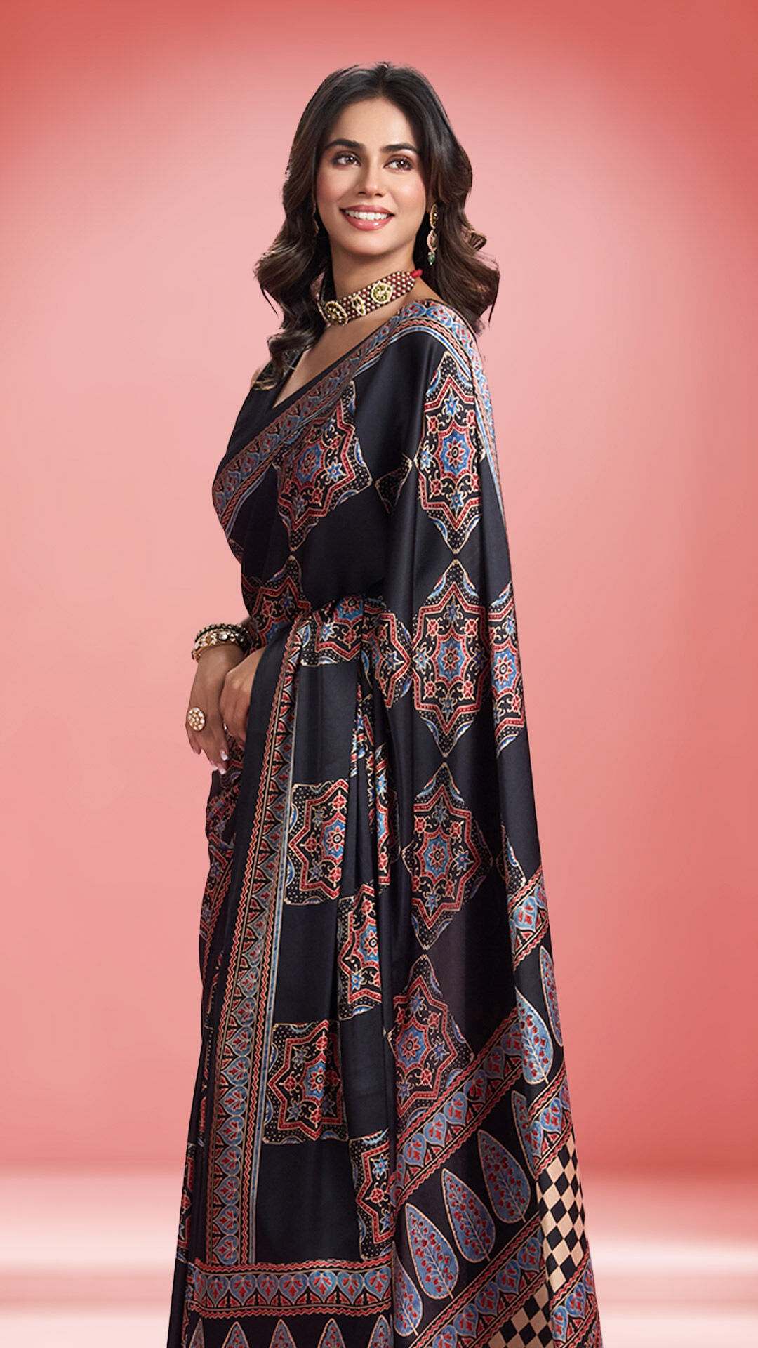 Woman wearing a black saree with intricate red and blue ethnic motifs, styled with statement jewelry, standing against a blush pink backdrop.